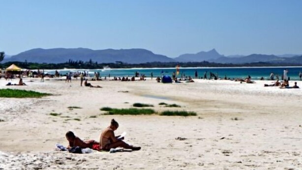 A beach with people sunbathing and swimming, a clear sky, and mountains in the background. - Main Beach Apartments (Byron Bay)