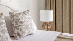 A hotel room with a white brick wall, a beige bedspread, floral-patterned pillows, a white lamp, and beige curtains. - Main Beach Apartments (Byron Bay)
