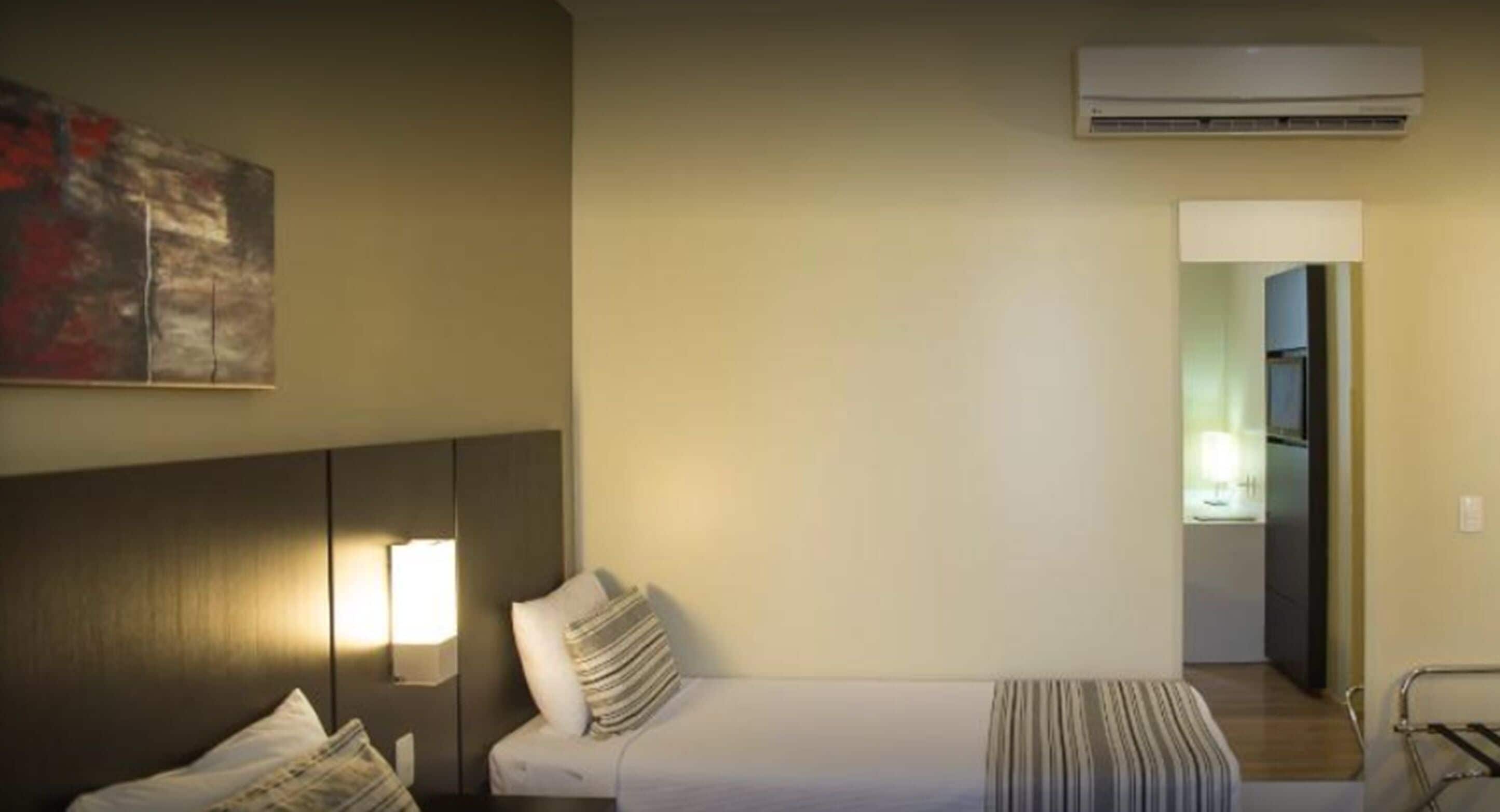 A hotel room with a bed, a nightstand, a lamp, a painting on the wall, and an air conditioning unit.