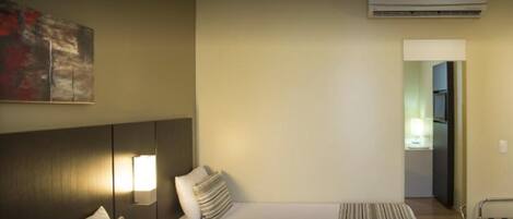 Una habitación de hotel con cama, mesita de noche, lámpara, un cuadro en la pared y un equipo de aire acondicionado.