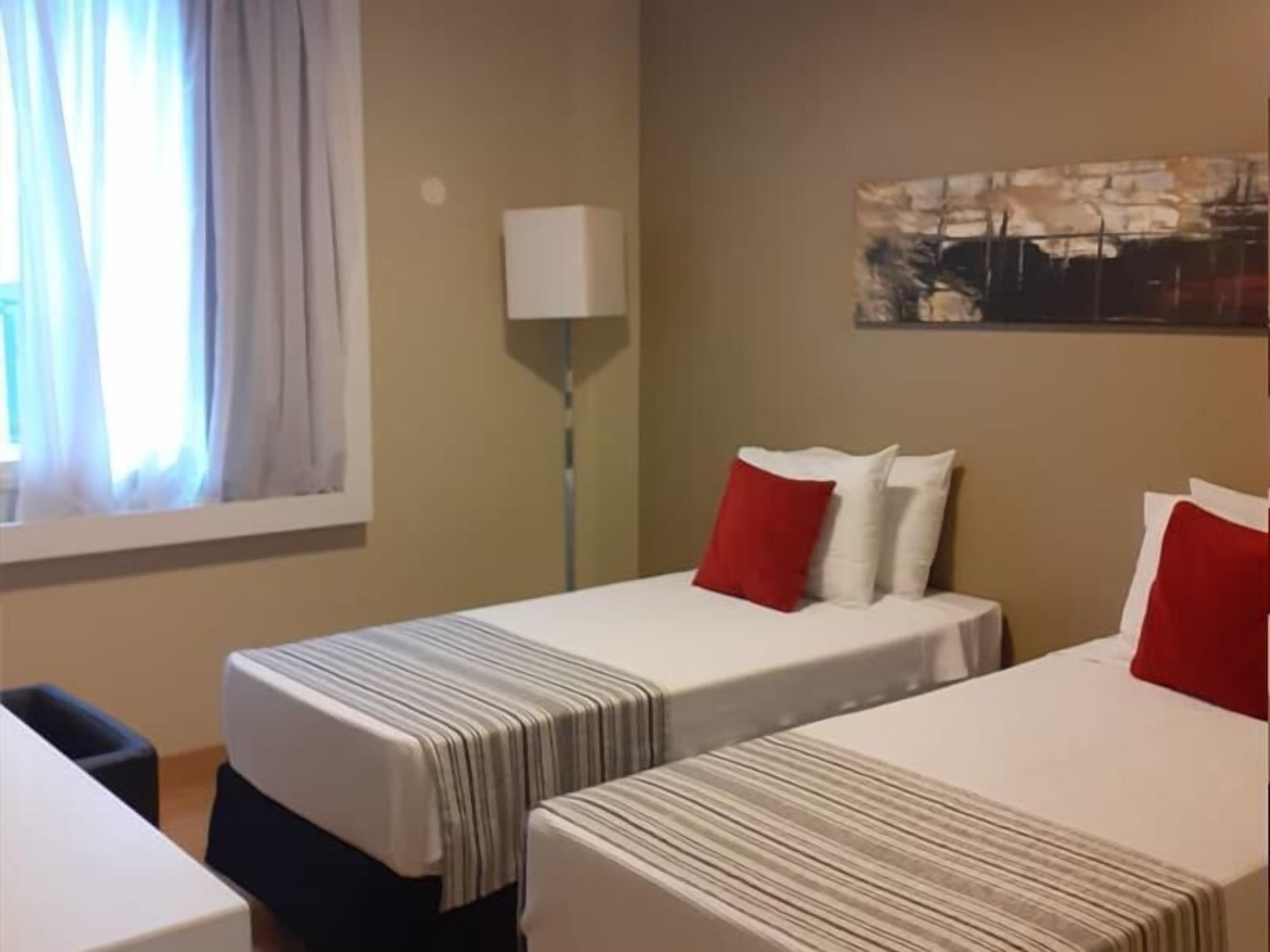 Habitación de hotel con dos camas, una ventana con cortinas, una lámpara y un cuadro en la pared.