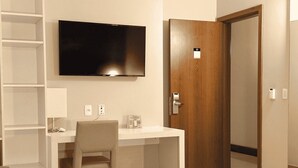 Habitación de hotel con cama, escritorio y silla, televisor montado en la pared y puerta de madera.