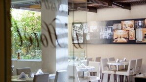 Un restaurante con mesas y sillas dispuestas en el interior, un muro de cristal con vistas a la vegetación exterior y una pared decorada con cuadros.