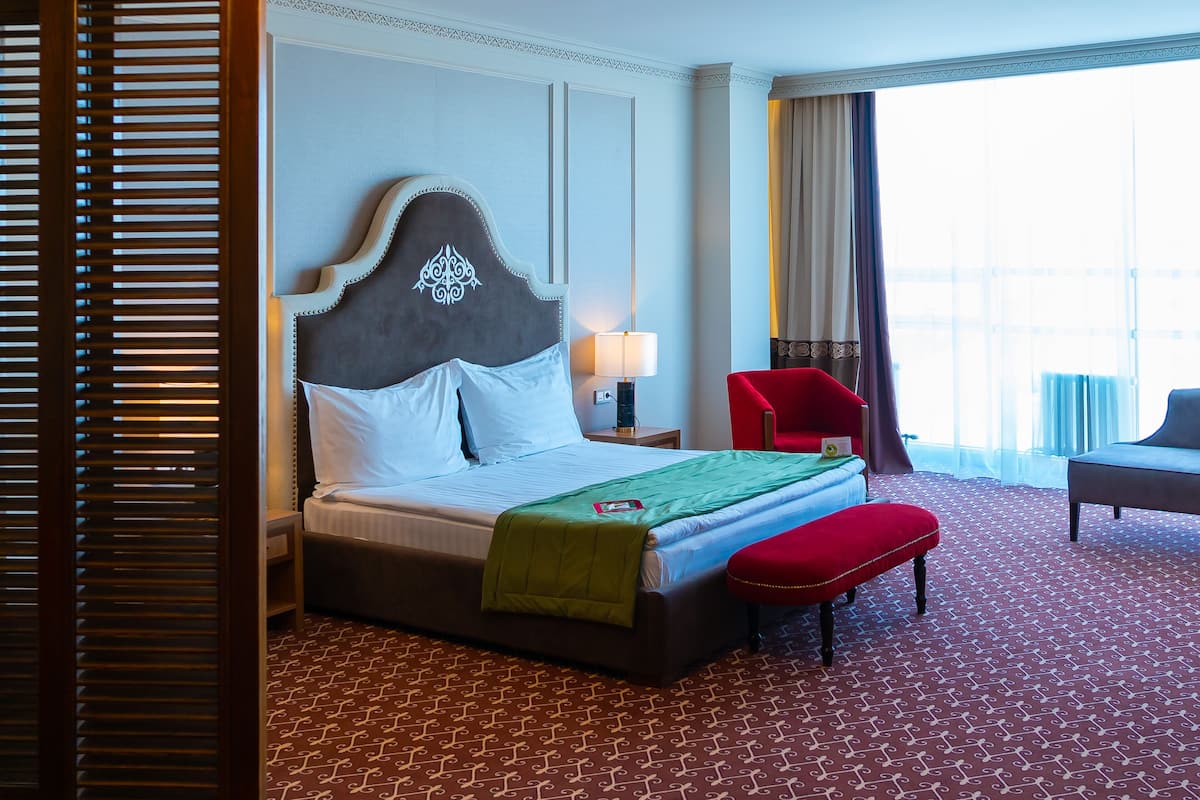 A hotel room with a large bed, a red armchair, a bench, a chandelier, and a view of the city.
