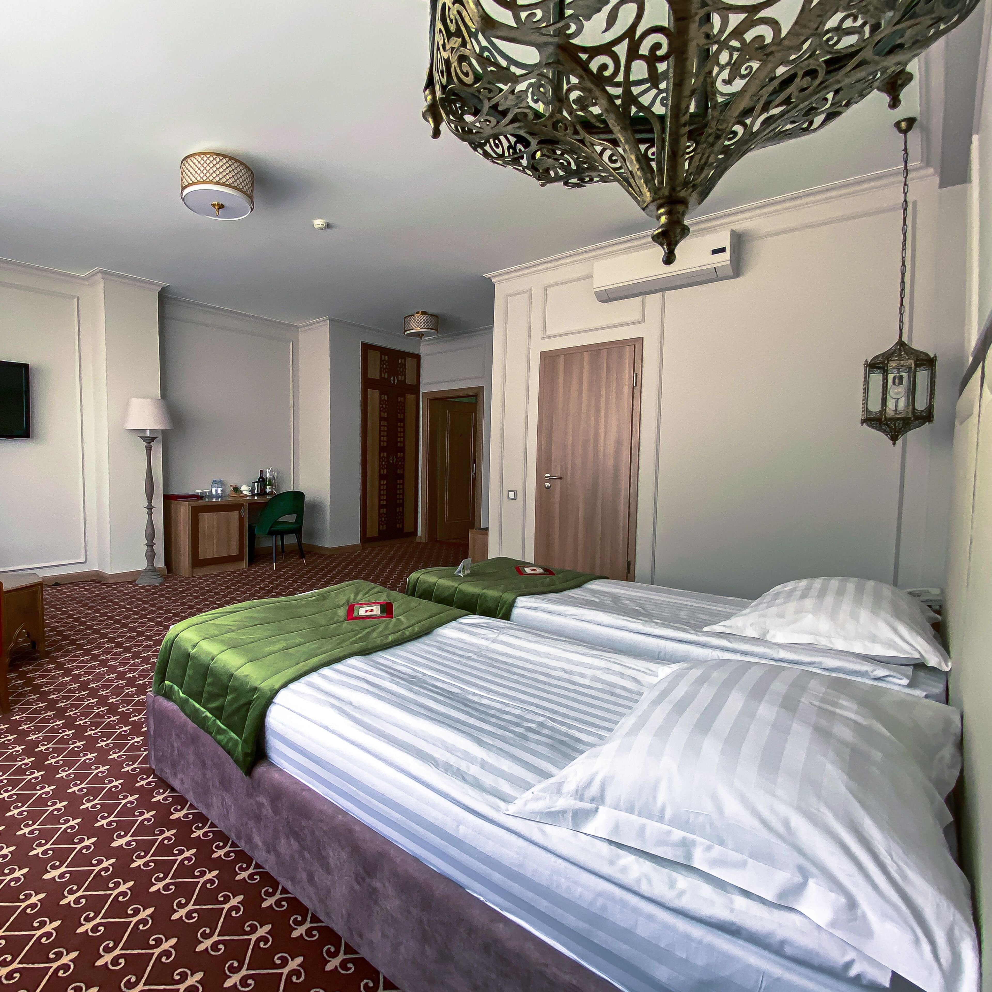 A hotel room with two beds, a desk, a chair, a wardrobe, and a decorative ceiling light fixture.