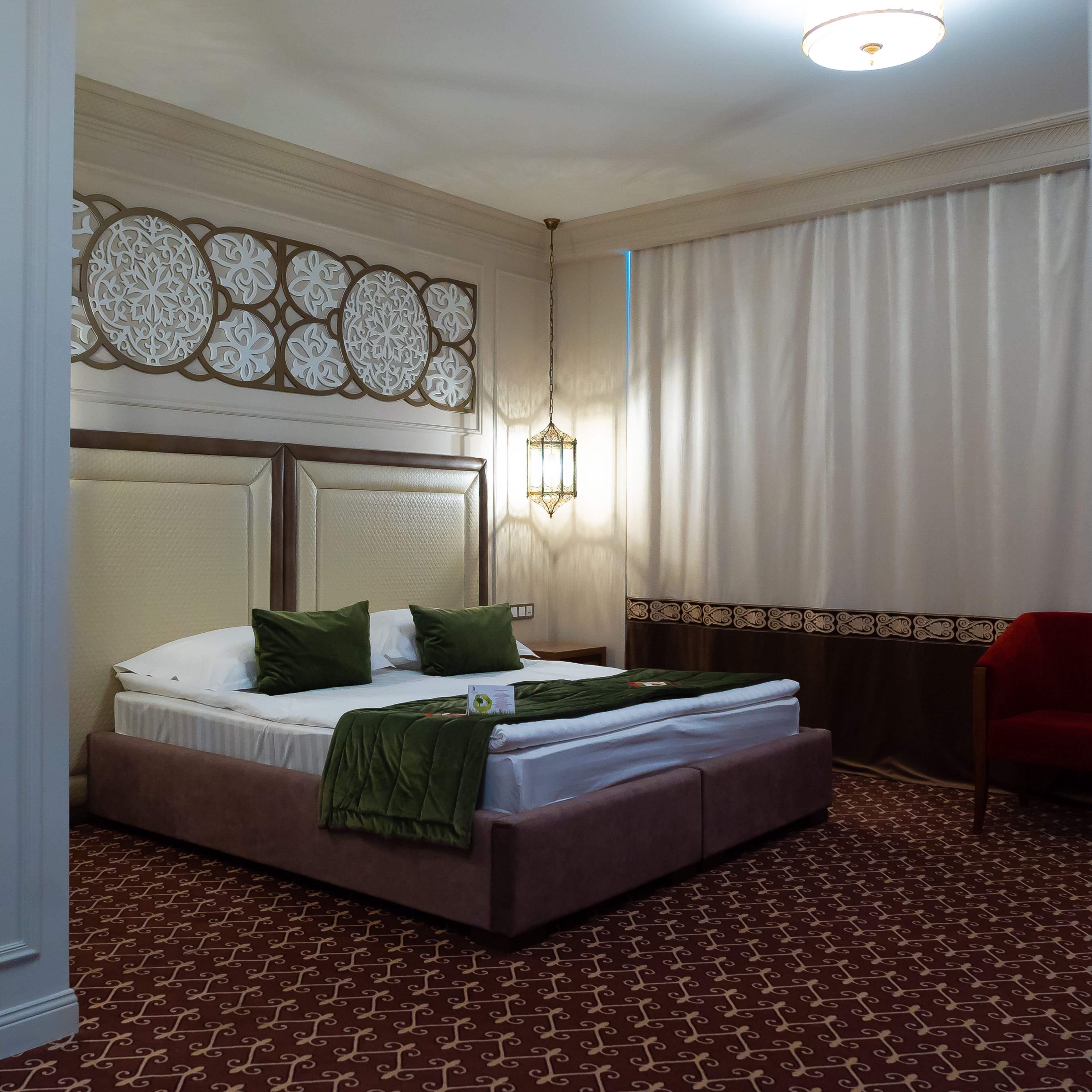 A hotel room with a bed, green pillows, a headboard, a nightstand, a red chair, and a patterned carpet.