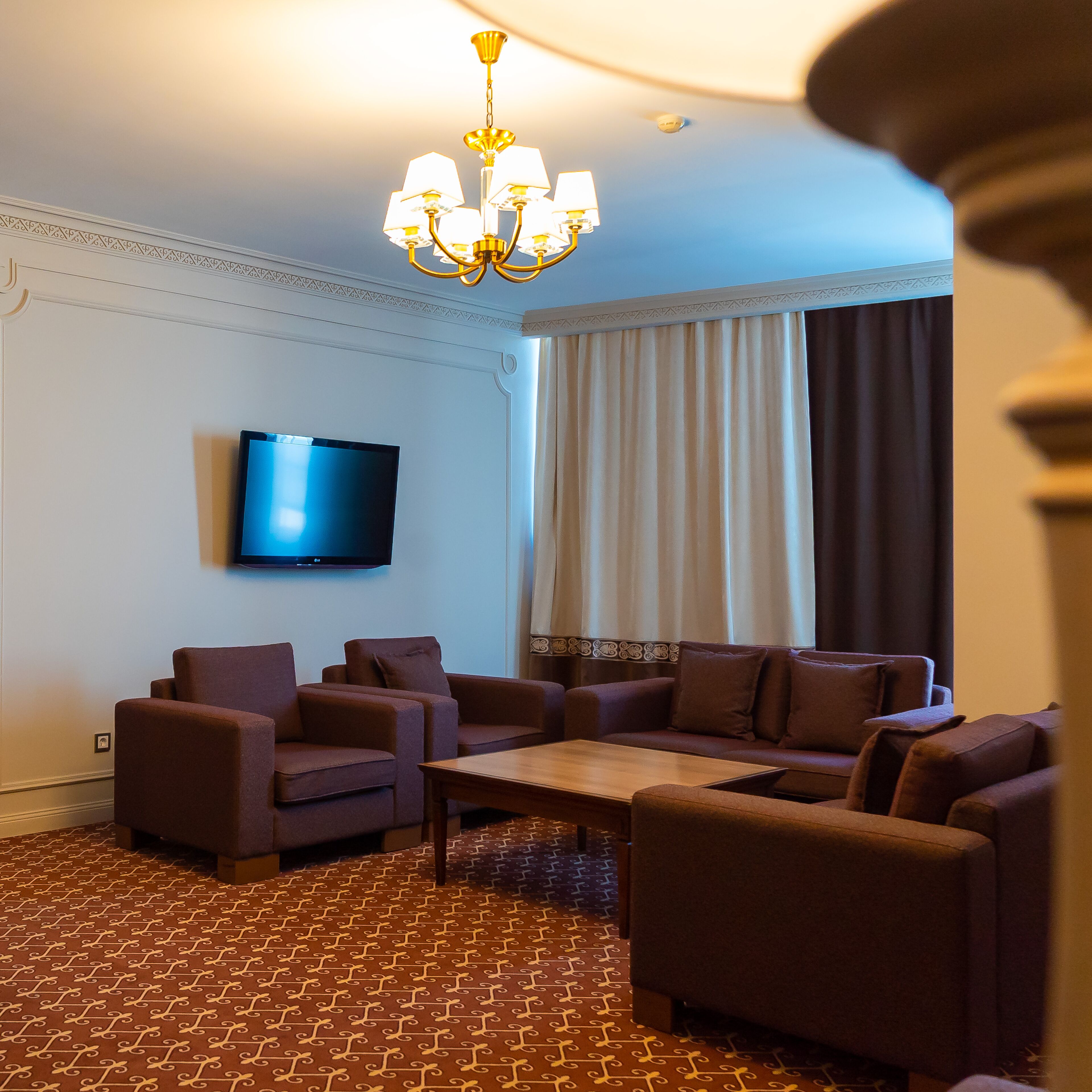 Habitación de hotel con televisión de pantalla plana montada en la pared, alfombra marrón estampada, un conjunto de sofá marrón y una araña.