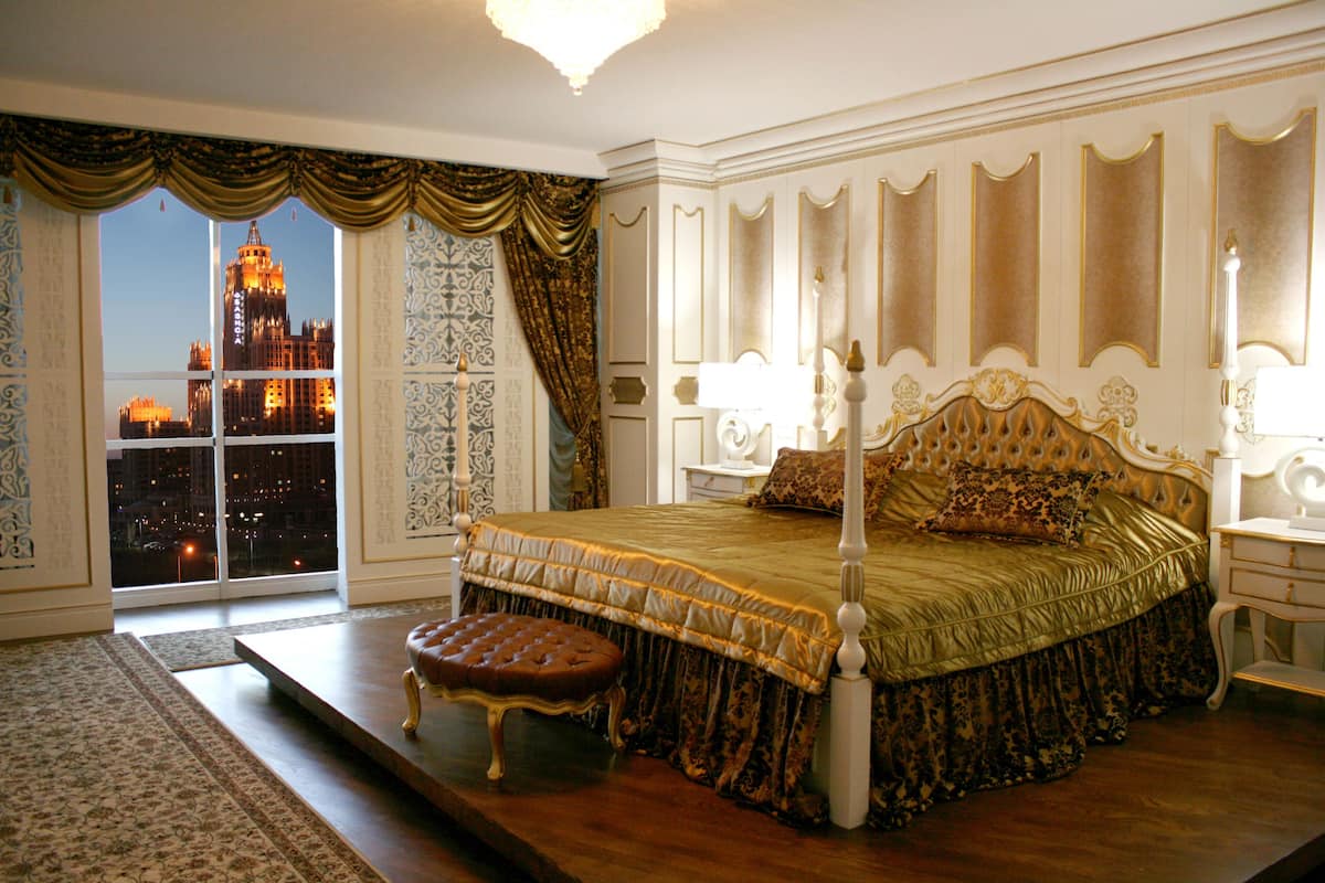 A luxurious bedroom with a four-poster bed, a nightstand with a lamp, and a view of a cityscape through a large window.