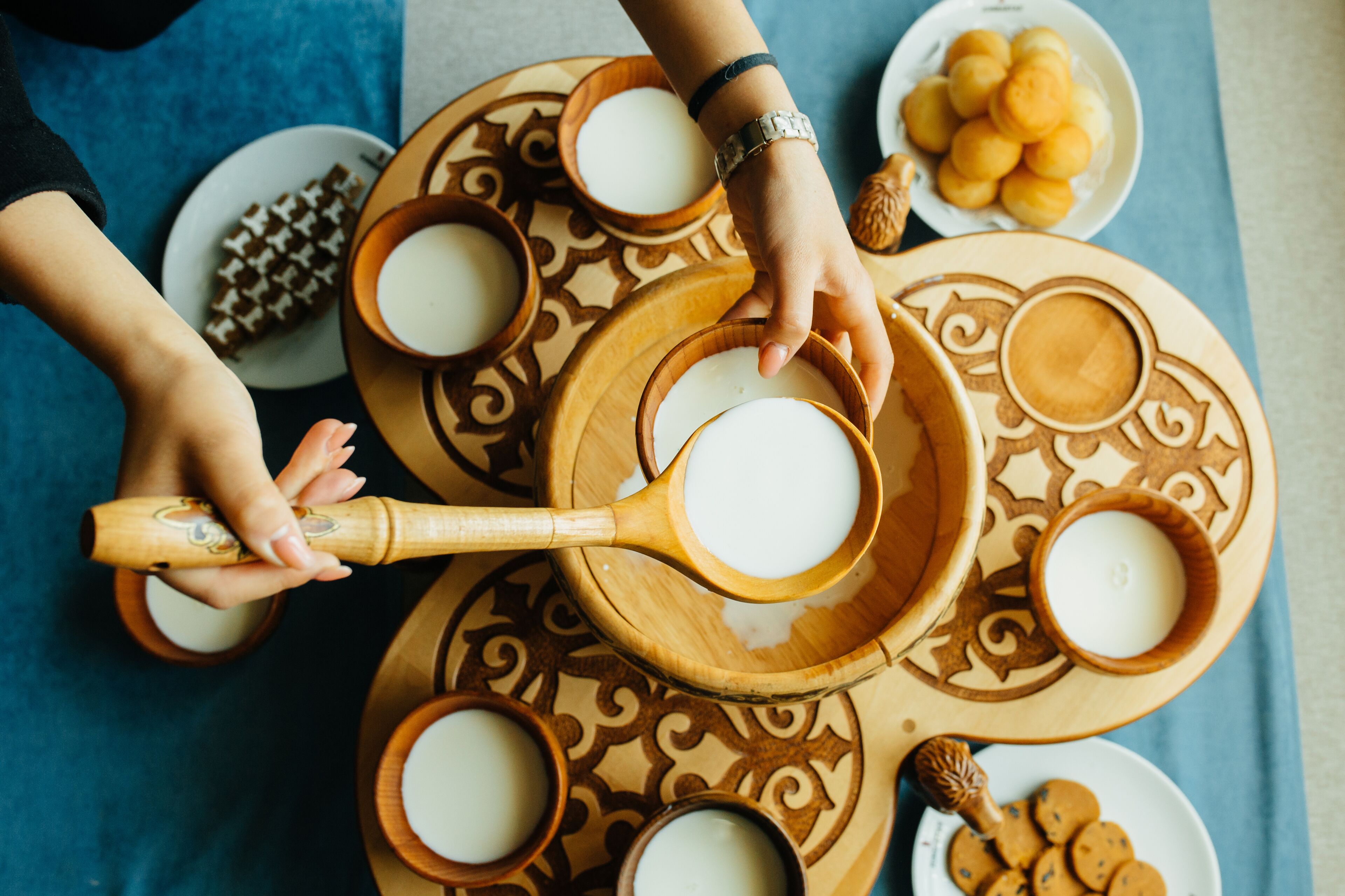 Eine Person gießt Milch in eine Holzschüssel mit langem Henkel, umgeben von anderen Schüsseln und Tellern mit einem dekorativen Muster.