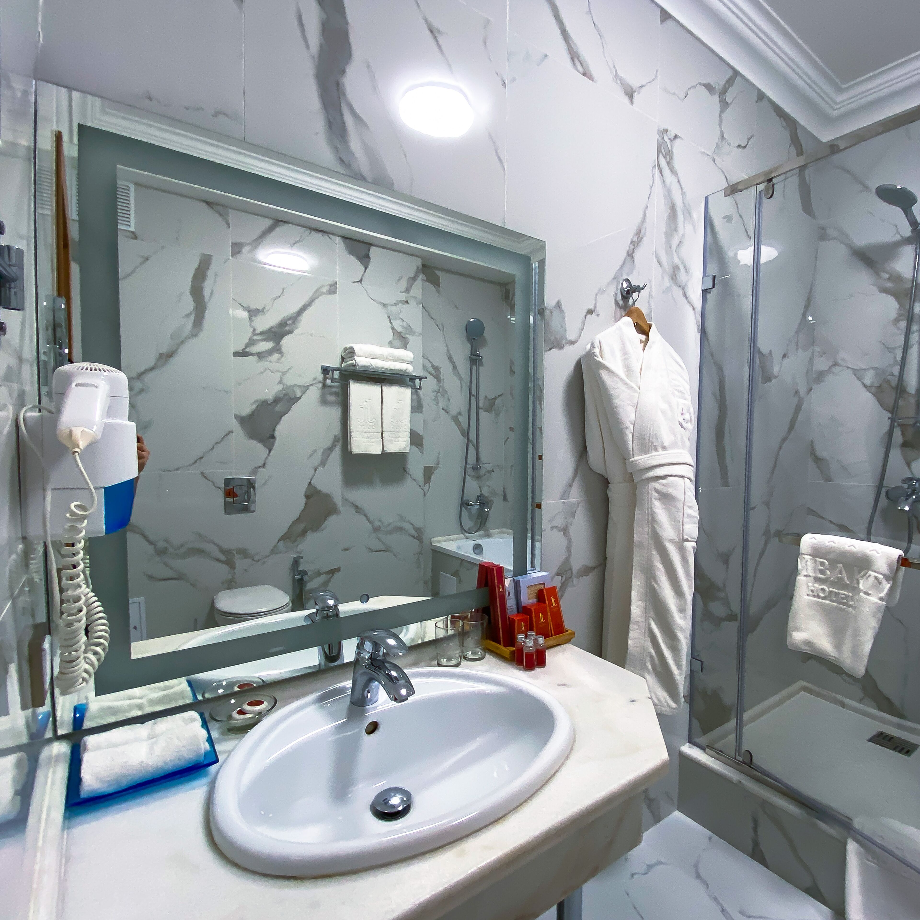 Een badkamer met een marmeren wand, een grote spiegel, een wastafel, een douchecabine van glas en een badjas aan de muur.