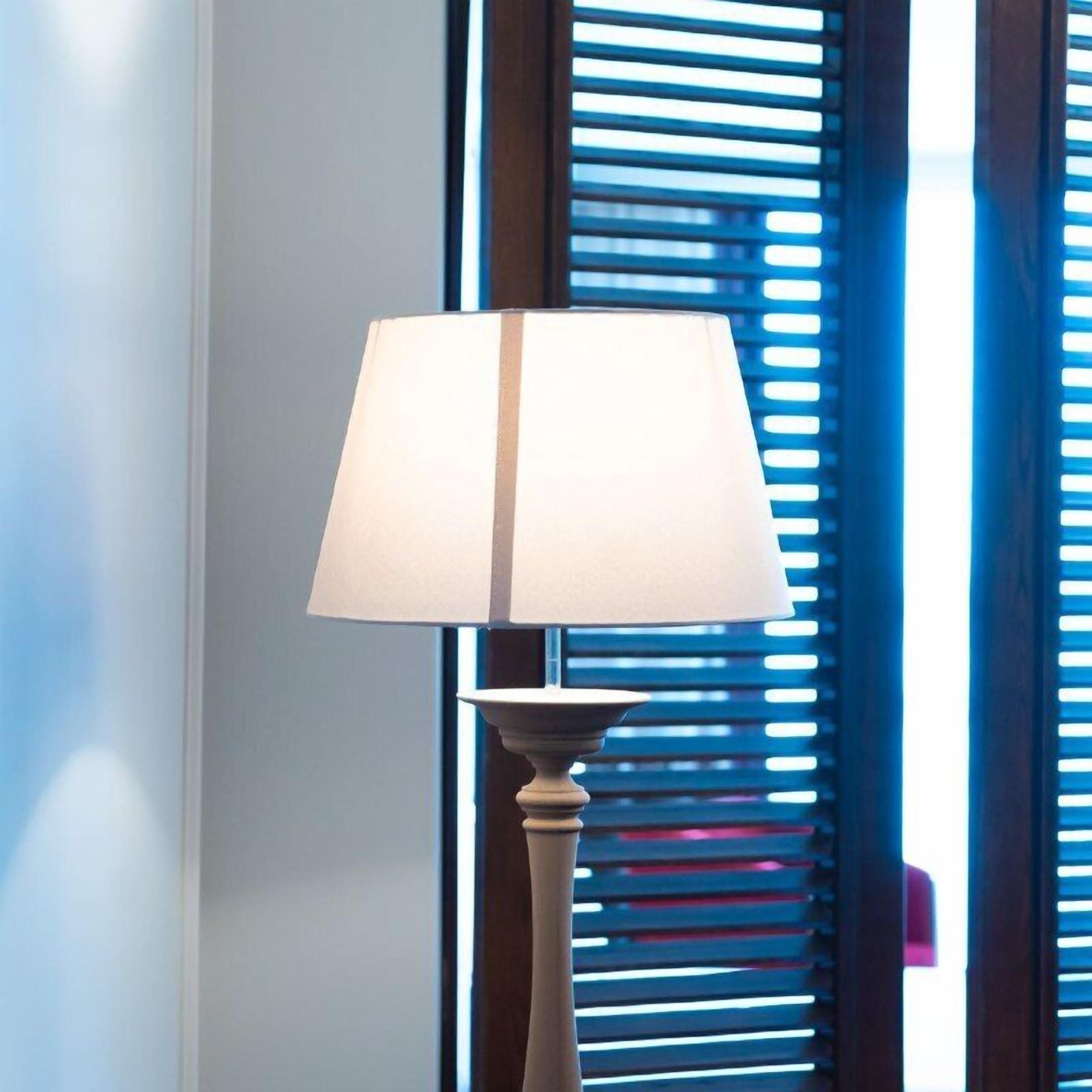 A lamp with a white shade on a pedestal, positioned in front of a window with horizontal blinds.