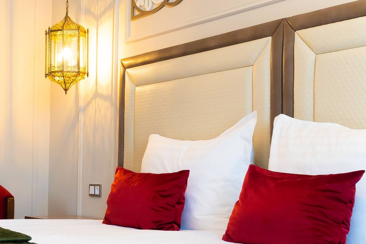 A hotel room with a large bed, white bedding, red and white pillows, a green throw blanket, and a wall-mounted light fixture.