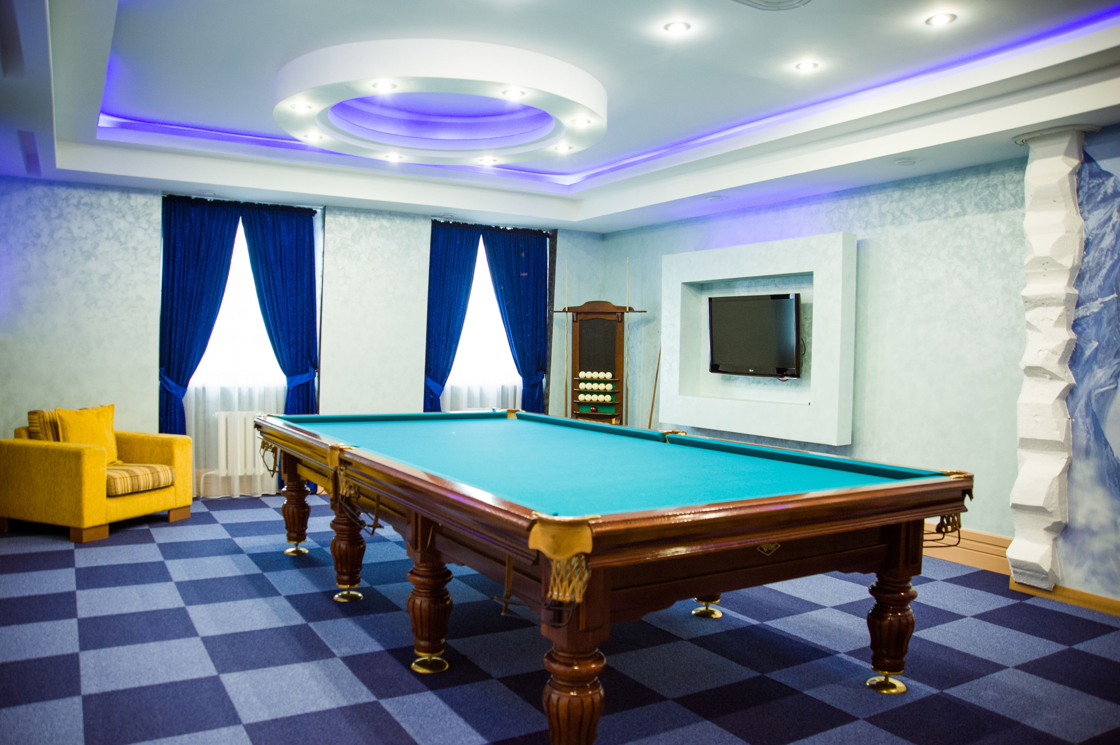 A pool table in a room with a checkered floor, a yellow armchair, and a television mounted on the wall.