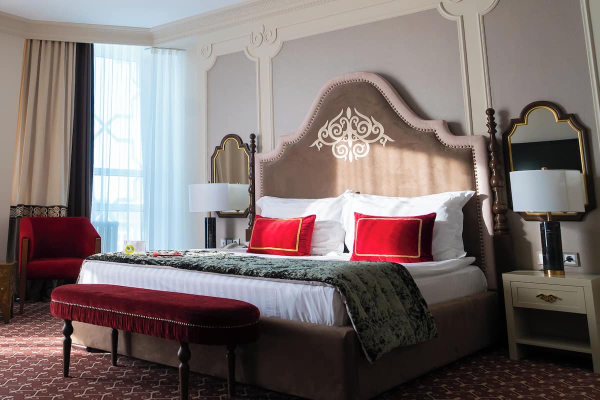 A hotel room with a large bed, two bedside tables, a red chair, a bench, a chandelier, and a large window with curtains.