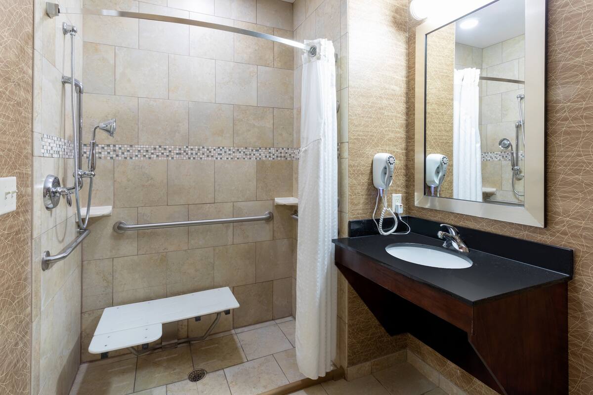 A bathroom with a shower, a sink, a towel dispenser, and a bench.