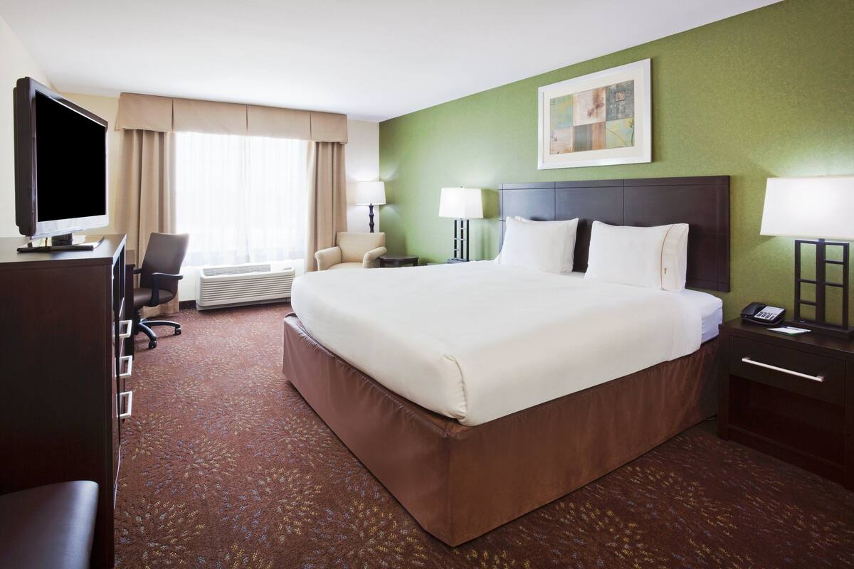 A hotel room with a large bed, a desk with a computer, a chair, a TV, and a window with curtains.