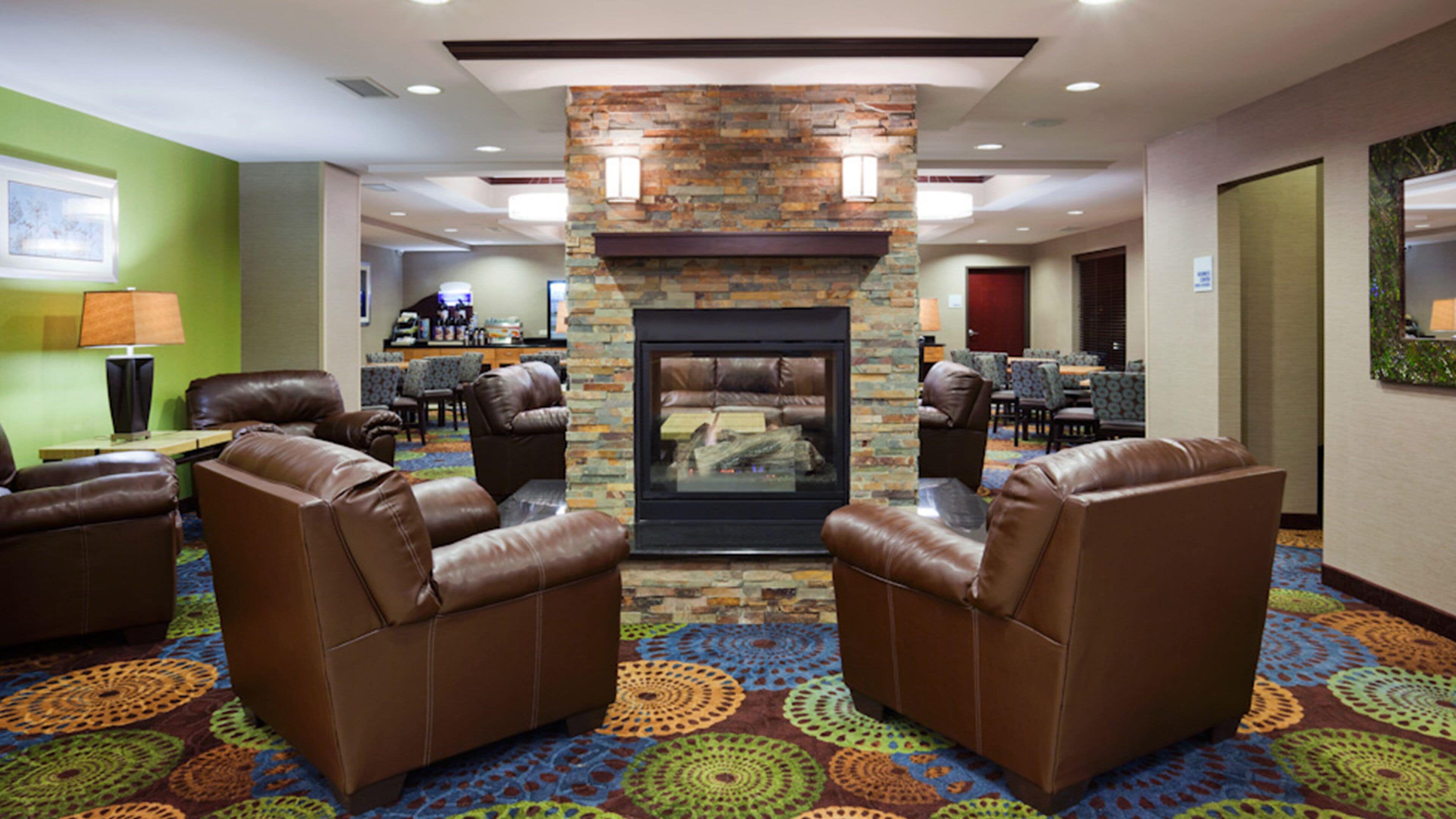 A hotel lobby with leather armchairs, a fireplace, patterned carpet, and a dining area in the background.