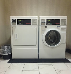 Two stacked commercial dryers in a laundry room with a tiled floor and a trash bin.