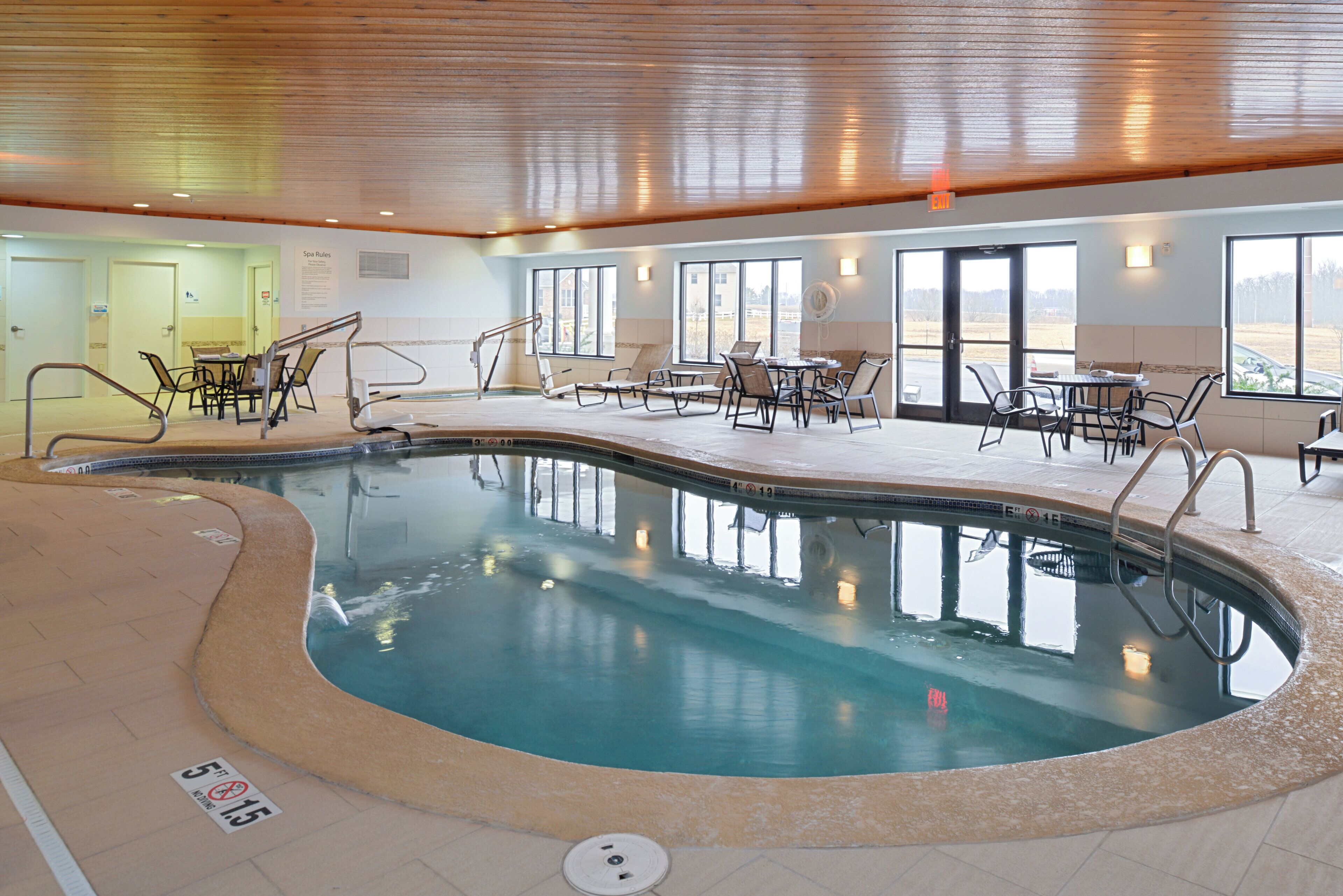 An indoor swimming pool with lounge chairs and tables, surrounded by windows and a wooden ceiling.
