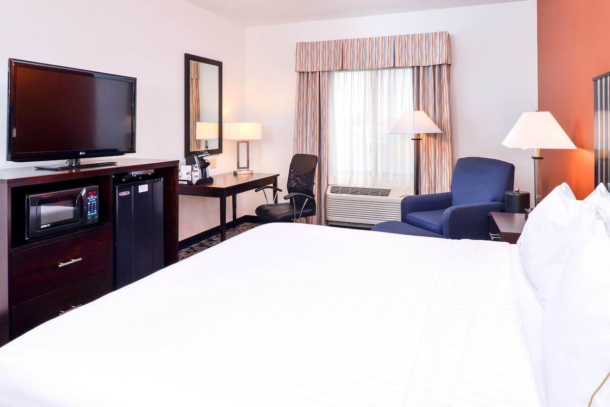 A hotel room with a large bed, a desk with a television, a chair, a desk lamp, a window with curtains, and an air conditioning unit.