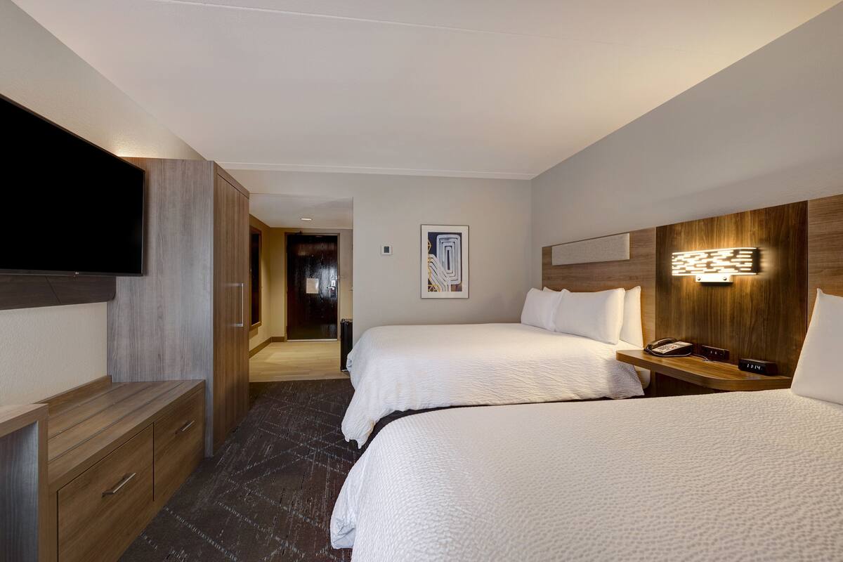 Une chambre d’hôtel avec deux lits, une télévision, une commode en bois, une table de chevet avec une lampe et un tableau au mur.