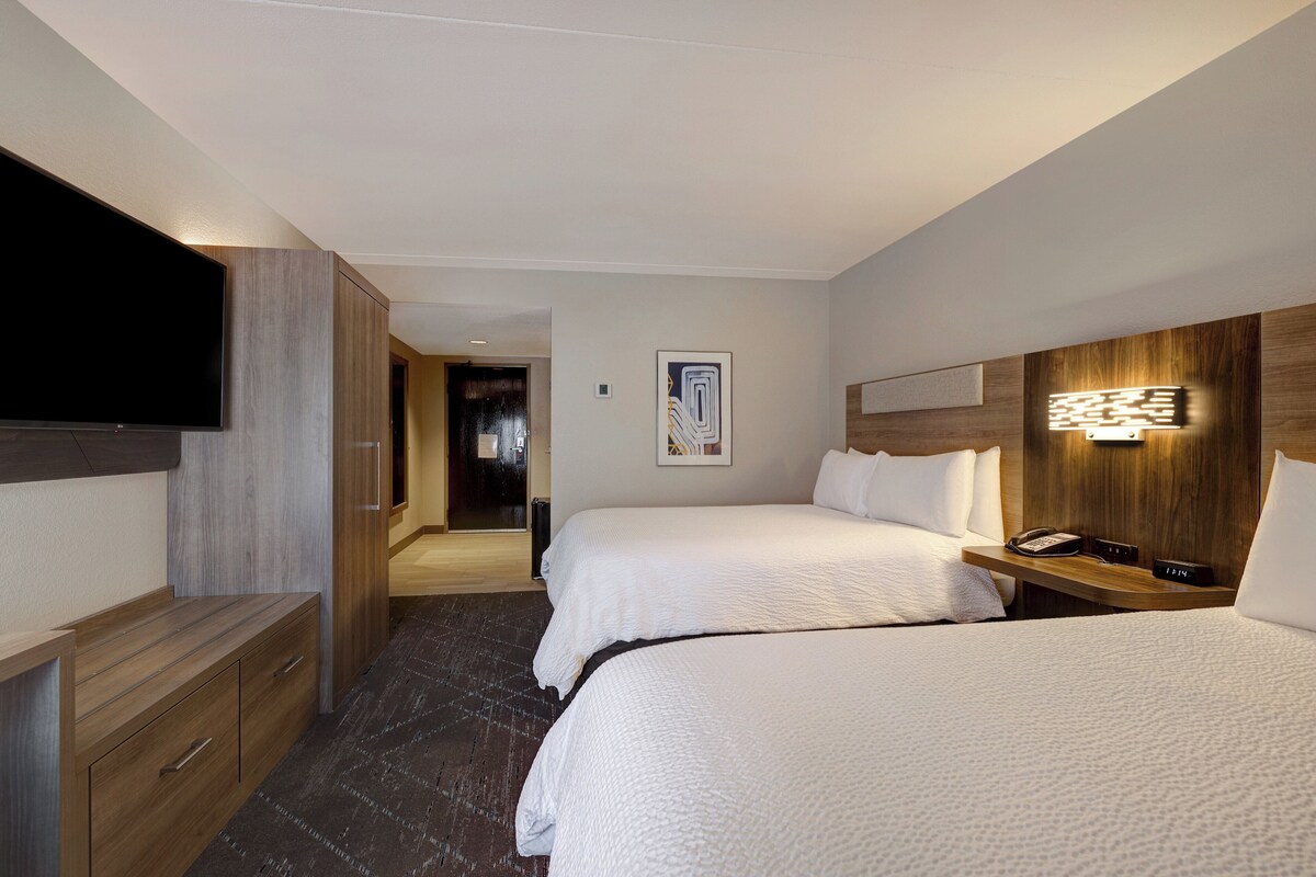 Een hotelkamer met twee bedden, een televisie, een houten dressoir, een nachtkastje met een lamp en een schilderij aan de muur.