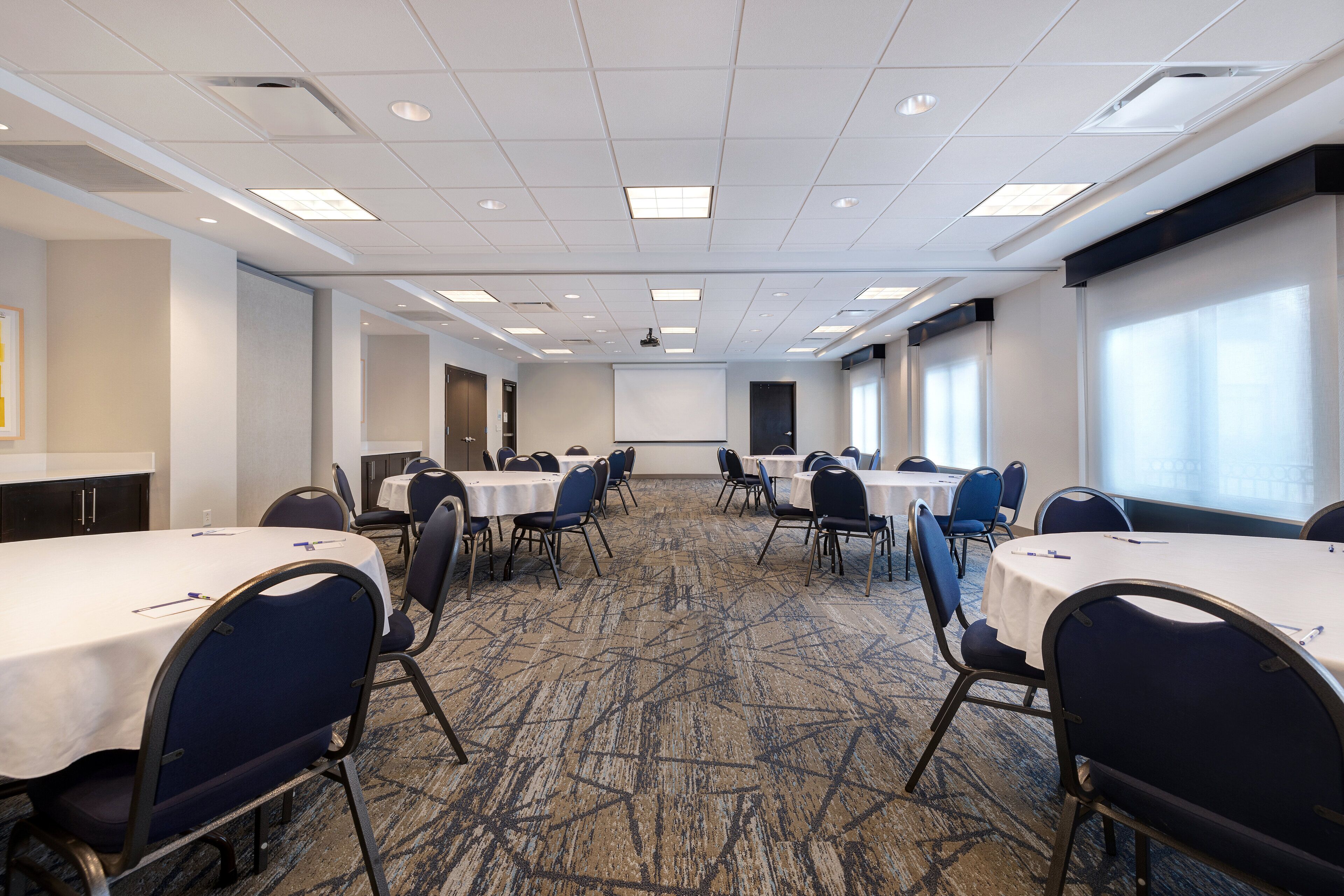 A conference room with multiple tables and chairs, a carpeted floor, and a projector screen.