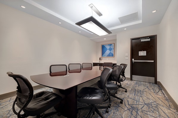 Une salle de conférence avec une longue table, quatre chaises, un sol recouvert de moquette, un luminaire au plafond et une porte avec une pancarte.