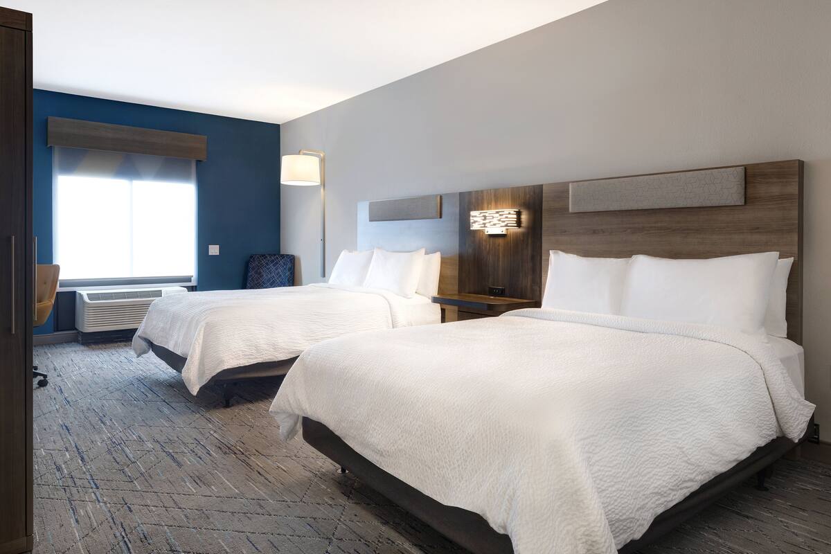 Une chambre d’hôtel avec deux lits, une grande fenêtre avec stores, un luminaire fixé au mur et une tête de lit en bois.