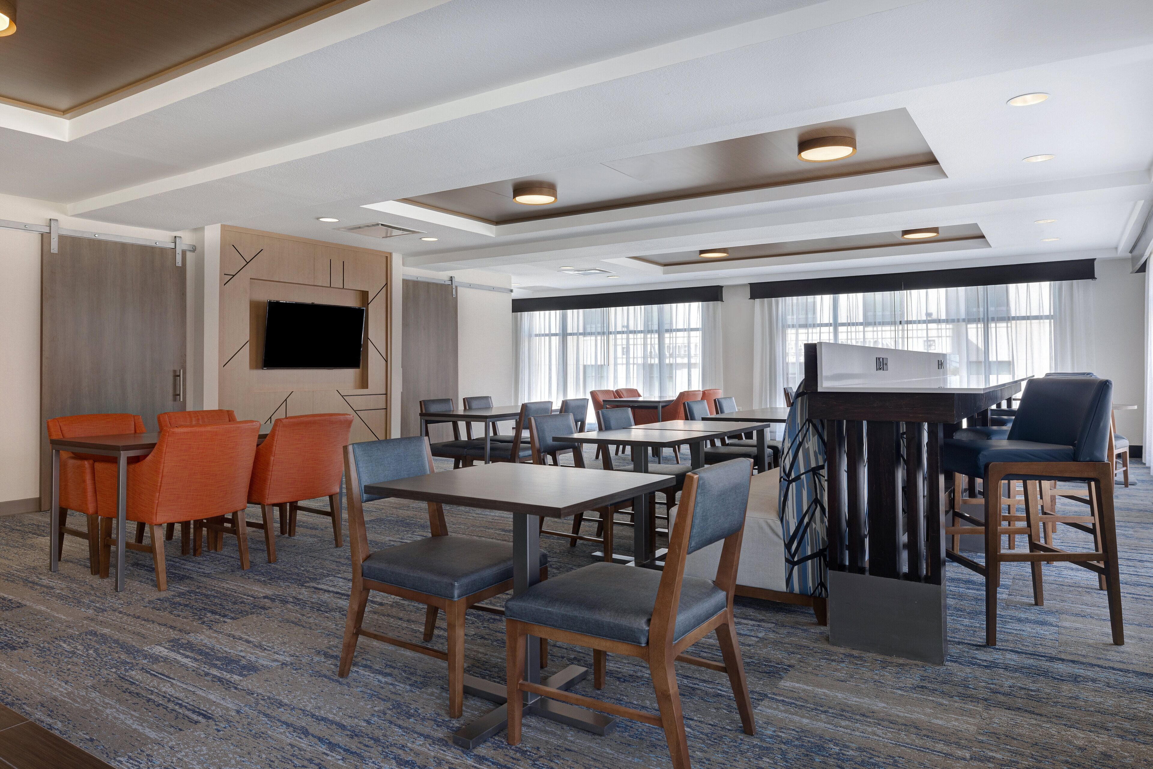 Um hall de hotel com várias mesas e cadeiras para refeições, uma TV de tela plana fixada na parede e um espaço de bar com balcão e banquetas.
