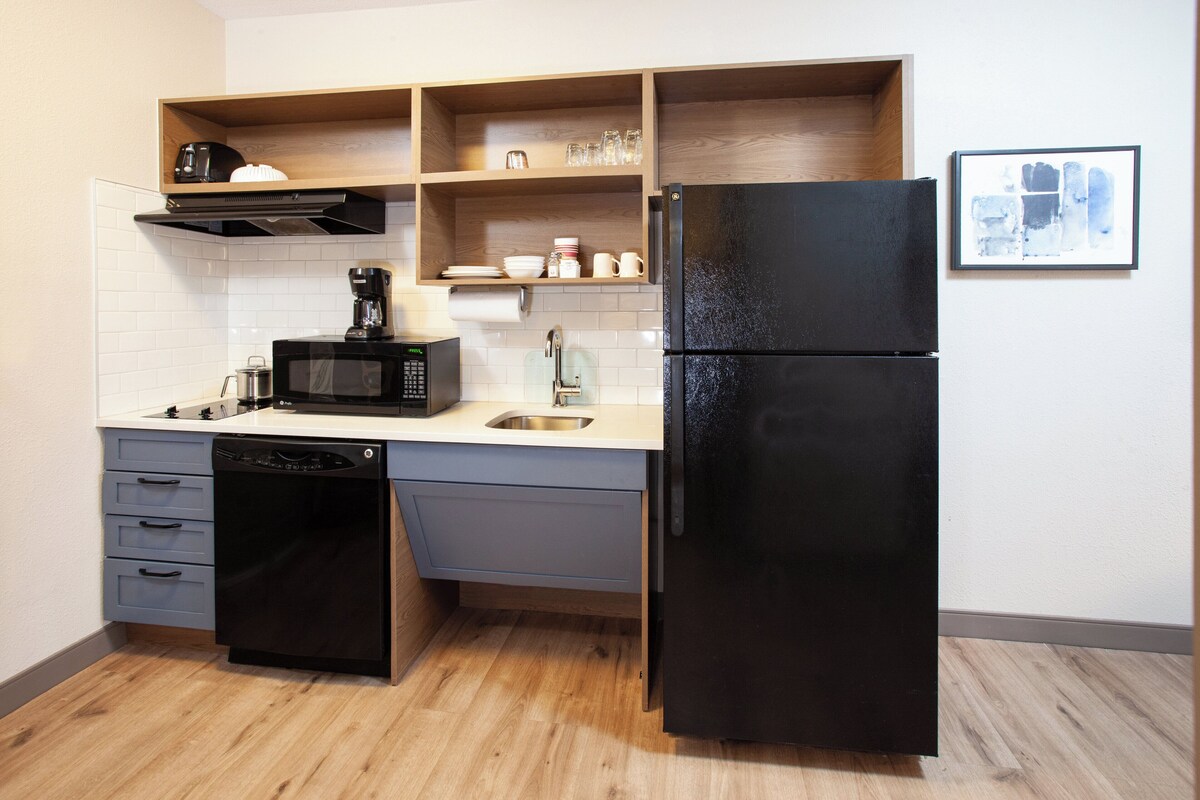 Cocina compacta con lavavajillas, microondas, refrigerador, estantes de madera y salpicadero de azulejos.