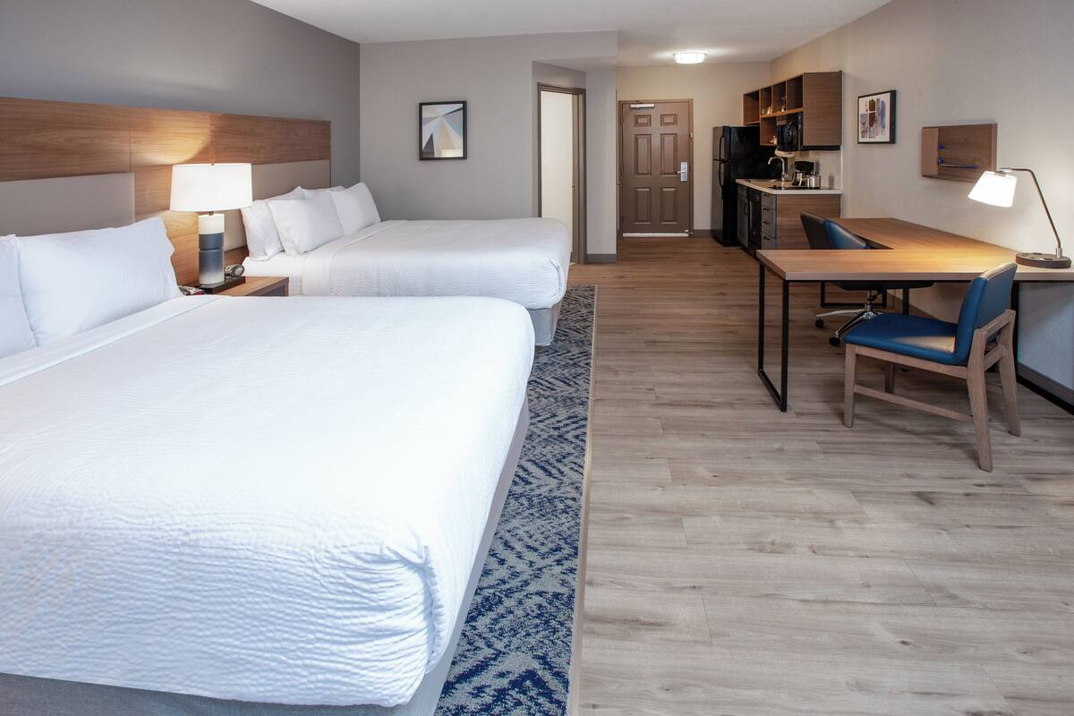 A hotel room with two beds, a desk with a chair, a kitchenette, and a door leading to another room.