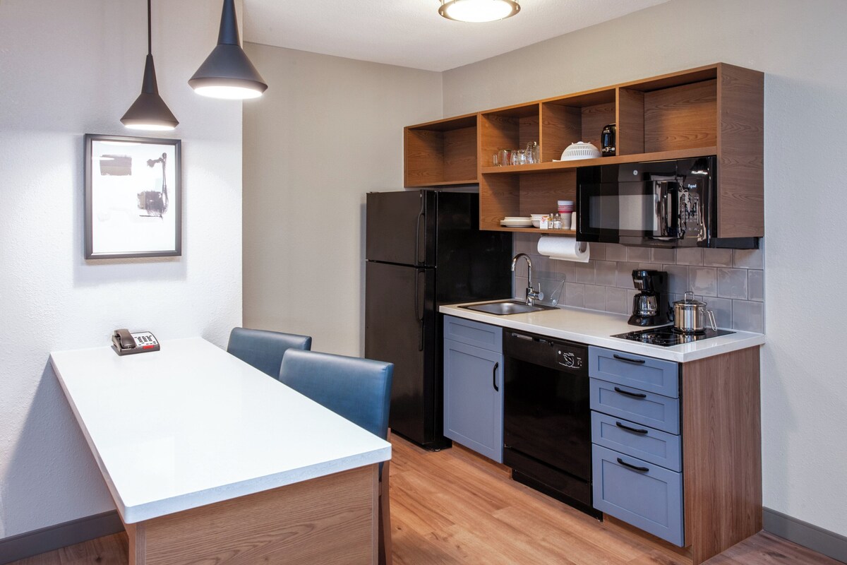 Cocina compacta con encimera blanca, electrodomésticos negros y estantes de madera.