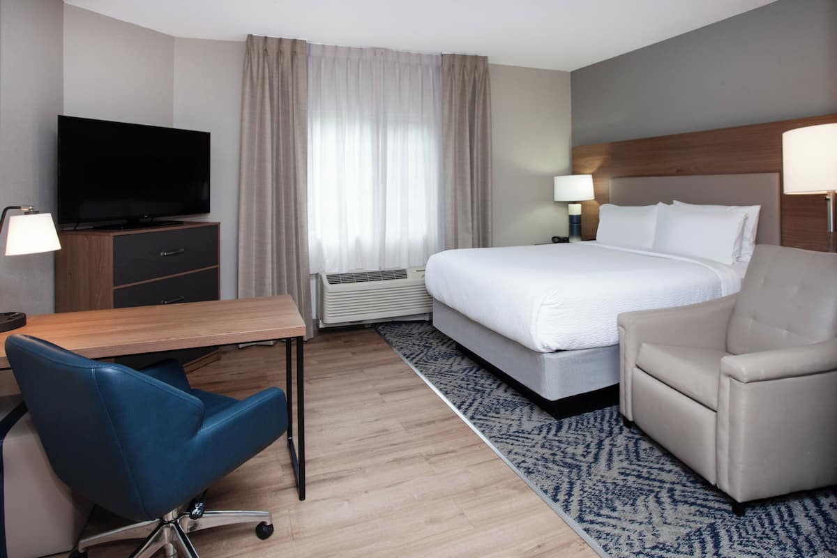 A hotel room with a bed, a desk with a television, a chair, a lamp, and a window with curtains.