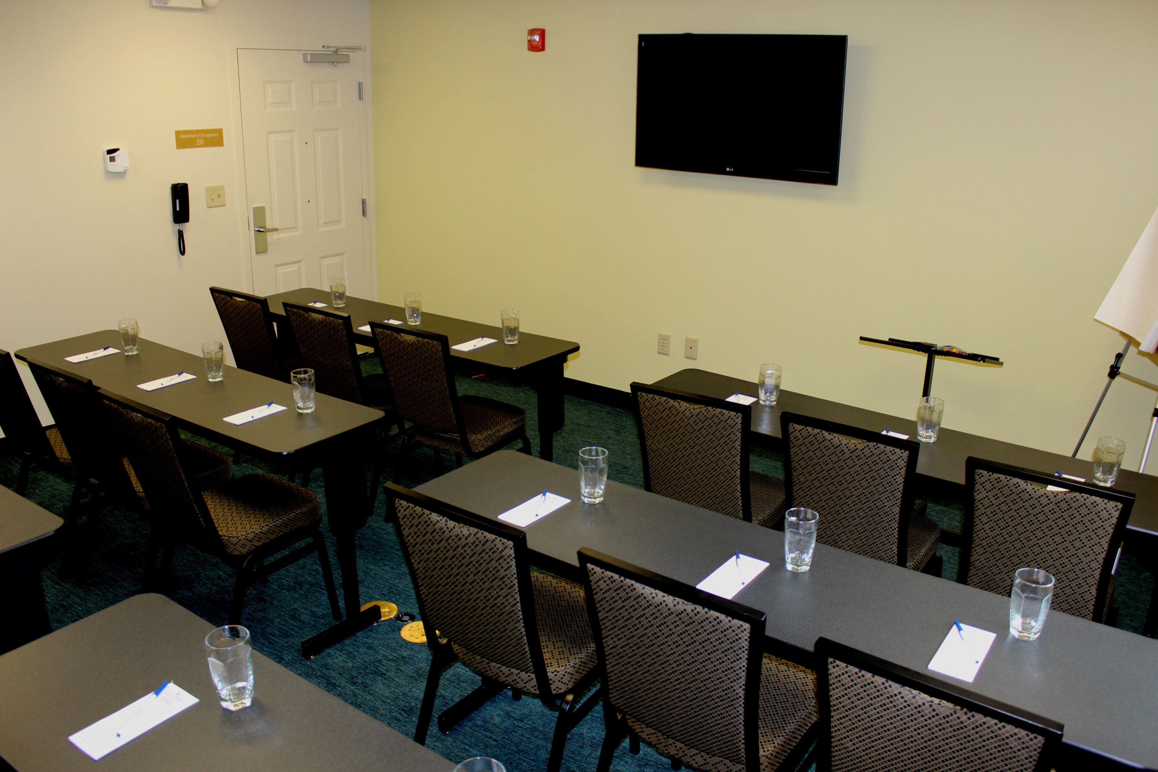 A conference room with tables, chairs, a flat-screen TV, and a door marked 'Maxim Room Occupancy 20'.