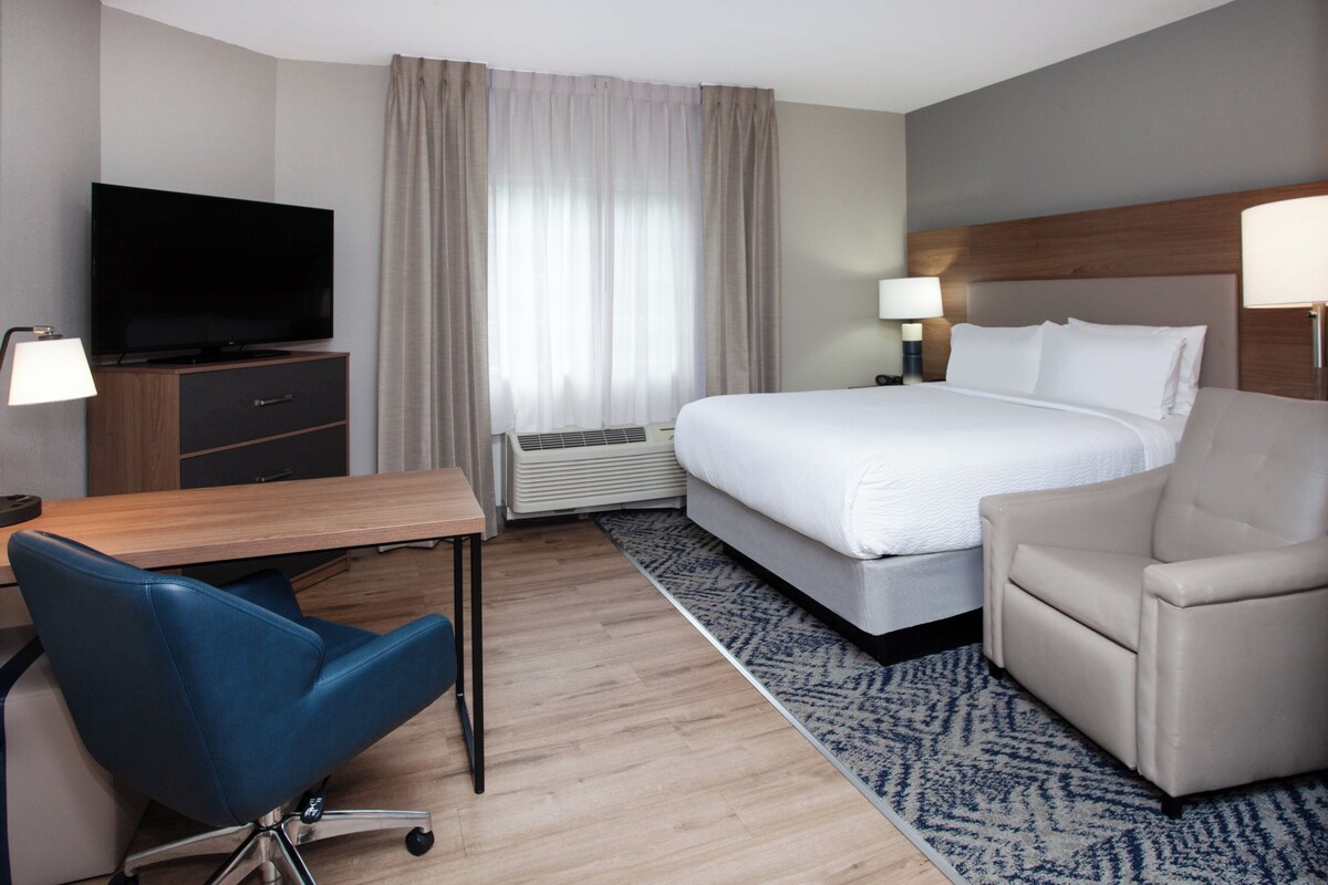 Habitación de hotel con cama, escritorio con televisor, silla, lámpara y ventana con cortinas.