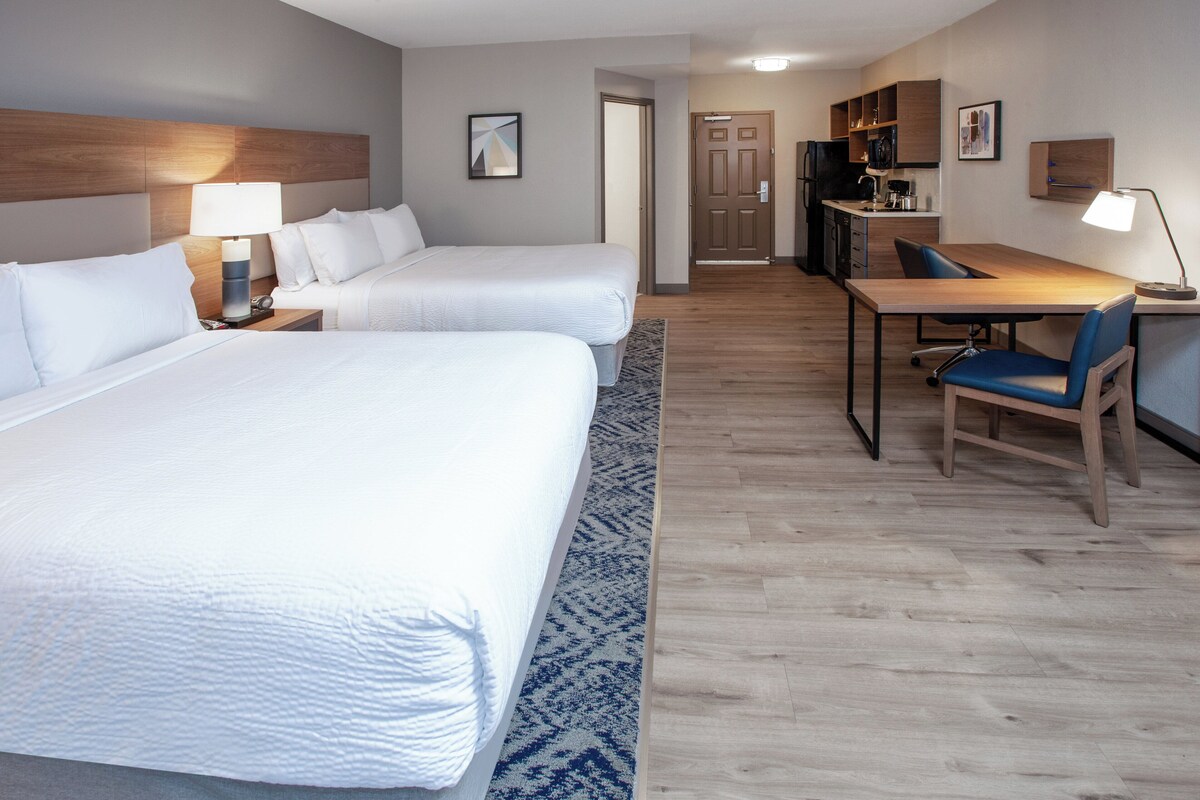 Una habitación de hotel con dos camas, un escritorio con una silla, una kitchenette y una puerta que da a otra habitación.