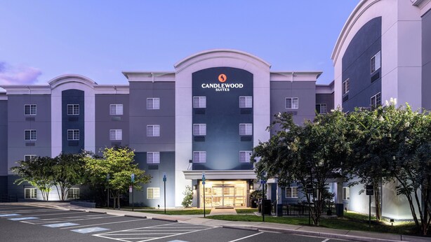 Un hotel de varios pisos con un llamativo letrero de "Candlewood Suites", un estacionamiento y un área ajardinada con árboles.