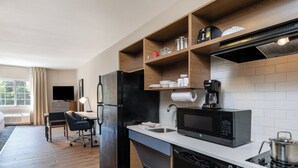 Una cocina moderna con refrigerador, microondas y lavavajillas, estantes de madera sobre la encimera y un comedor con mesa y sillas.