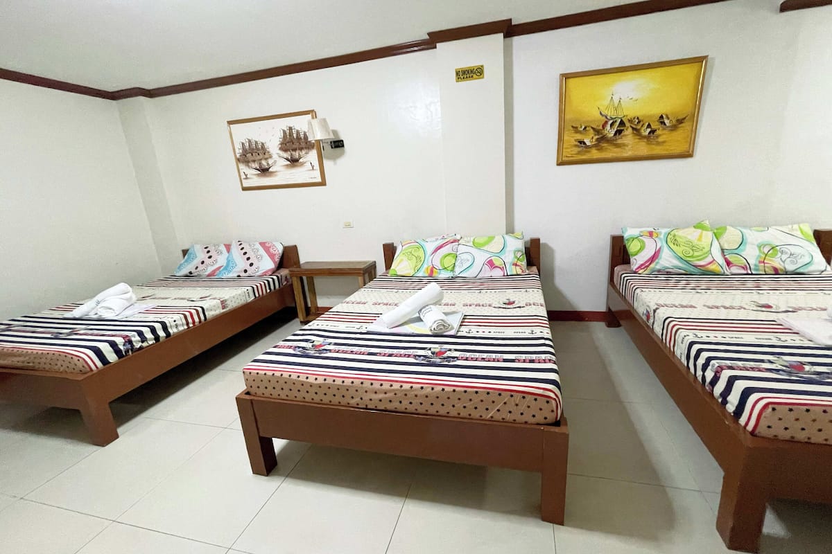 Une chambre d’hôtel avec trois lits simples, chacun recouvert d’une courtepointe à rayures et agrémenté d’oreillers assortis. Une petite table en bois se trouve entre les lits, et un tableau est accroché au mur au-dessus du lit de droite.