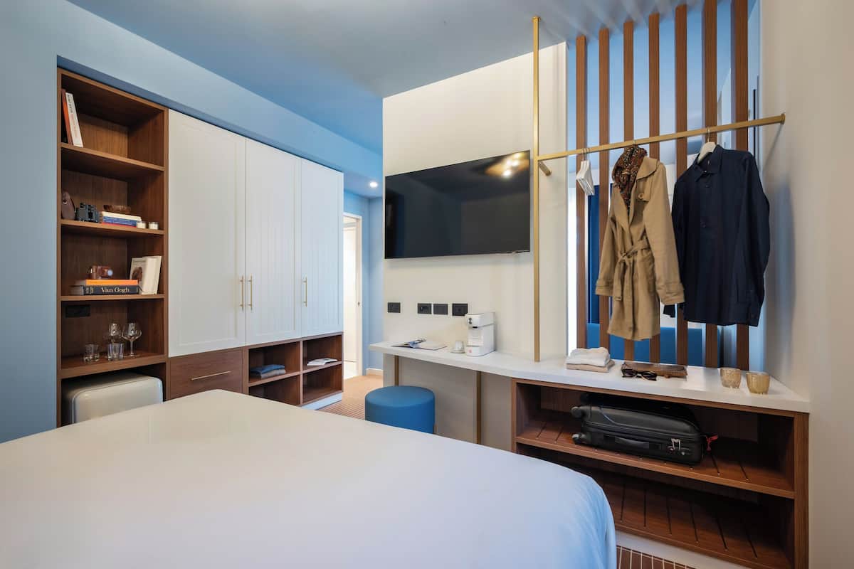 A modern hotel room with a bed, a desk with a flat-screen TV, a wardrobe, and a seating area with a blue ottoman.