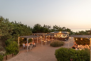 An outdoor patio area with tables and chairs, surrounded by greenery and a building with a sloped roof.