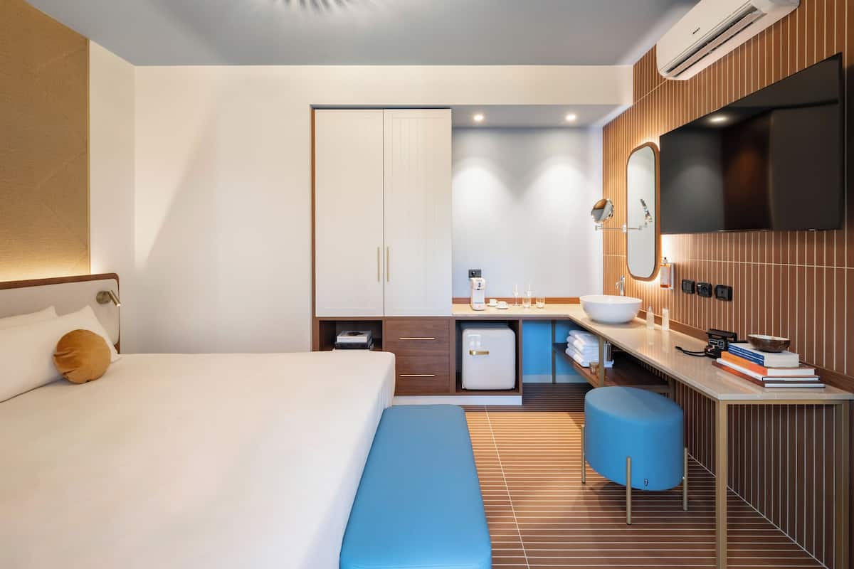 A modern hotel room with a bed, a desk with a computer, a sink, and a television.