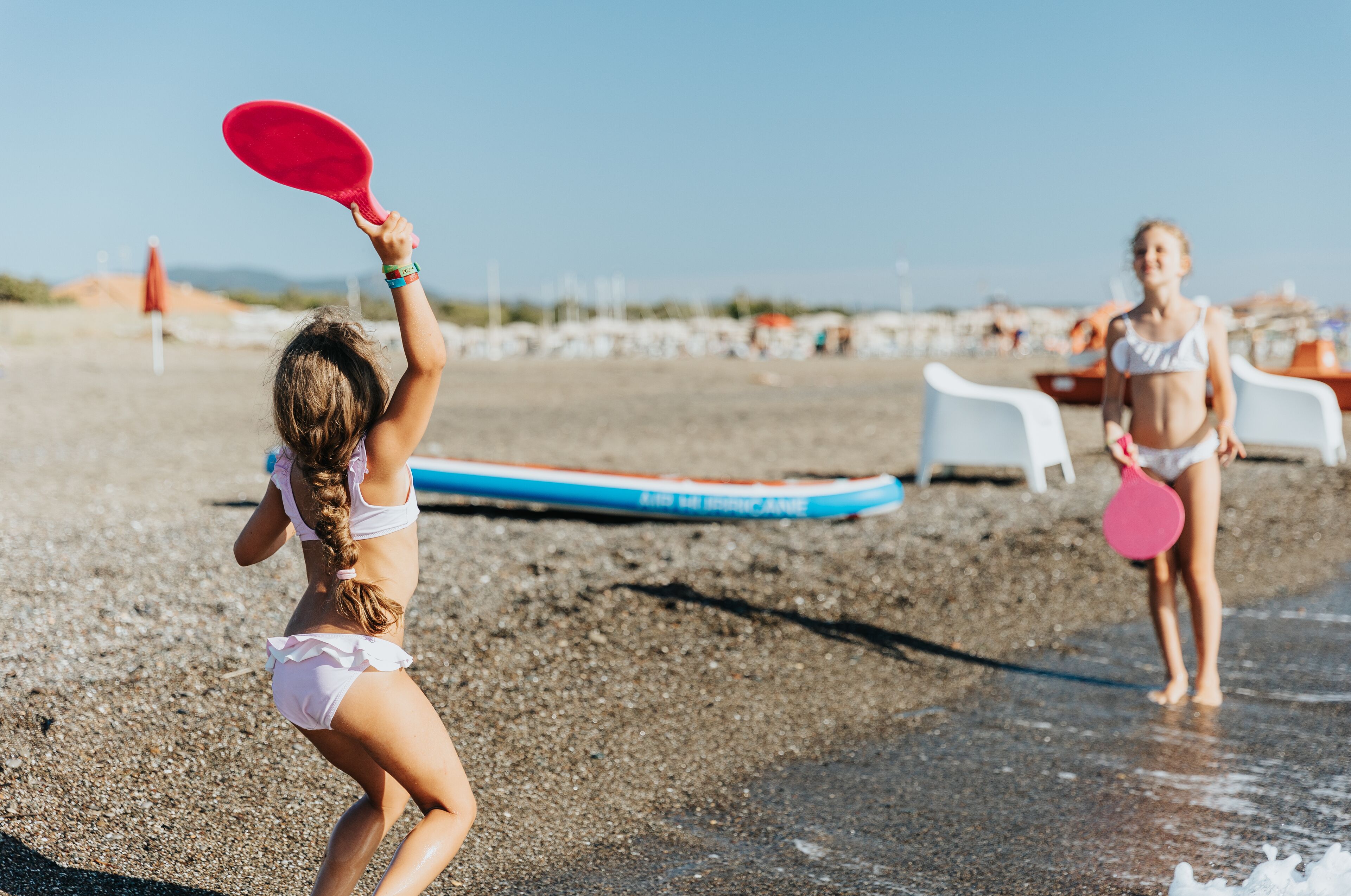 Una spiaggia con due bambini che giocano con pagaie, un’asse da surf blu e bianca e sedie da spiaggia bianche sullo sfondo.