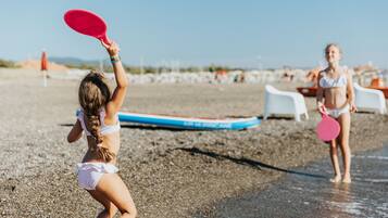 Una spiaggia con due bambini che giocano con pagaie, un’asse da surf blu e bianca e sedie da spiaggia bianche sullo sfondo.