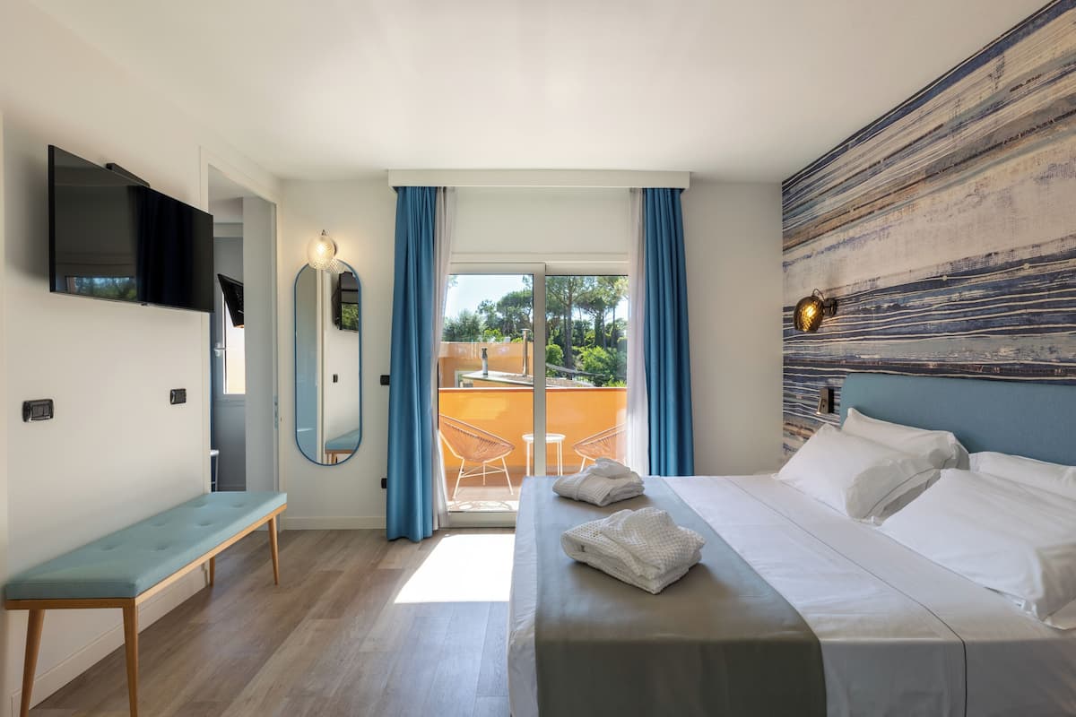 A modern hotel room with a bed, a bench, a TV, and a balcony with a view of the outdoors.