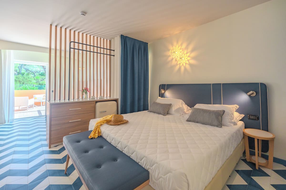 A bedroom with a bed, bedside table, bench, dresser, and a view of the outdoors.