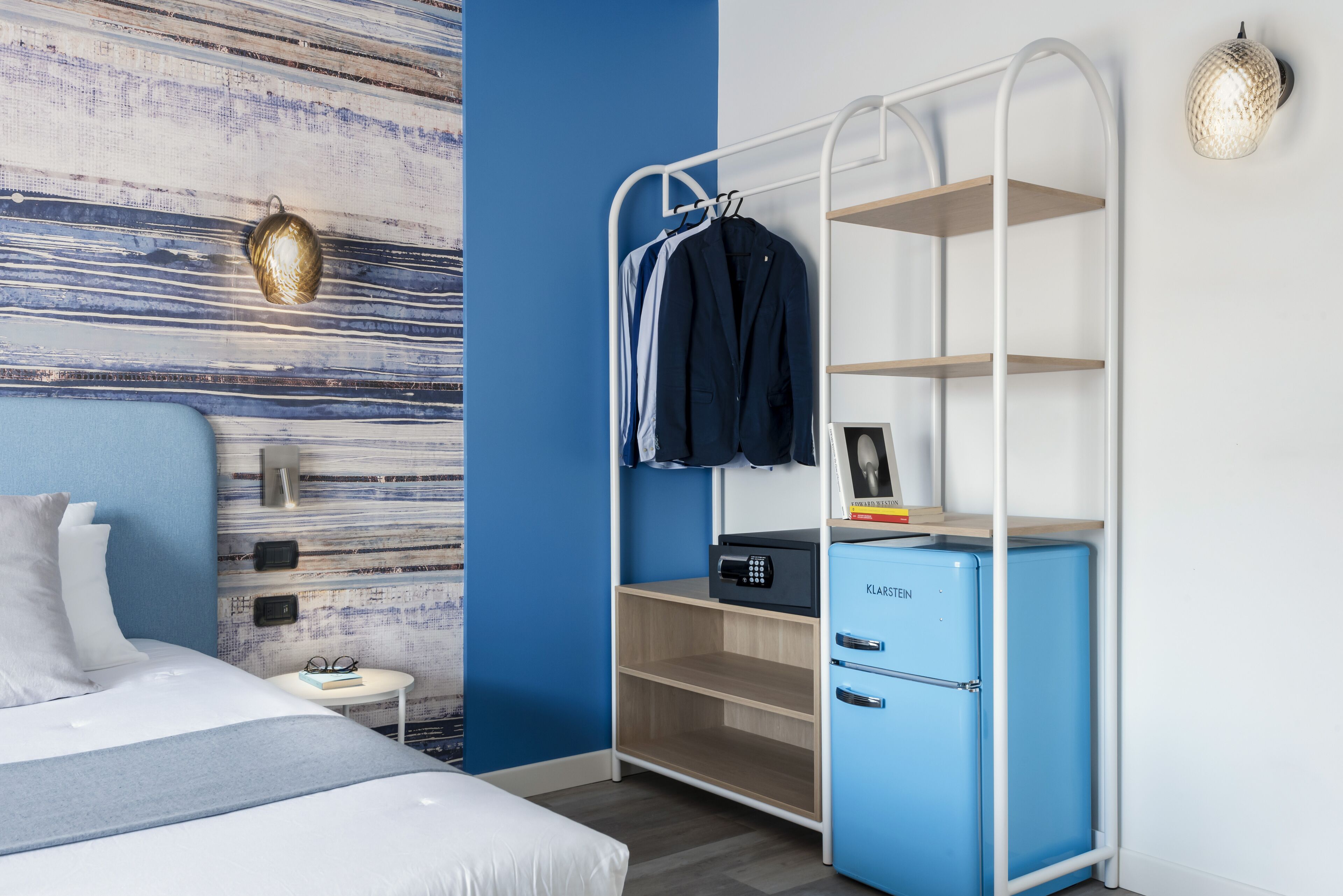 A modern hotel room with a bed, a small bedside table, a wall-mounted shelf with a safe, and a blue minibar.