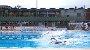 A swimming pool with lanes marked by floating lane dividers, a spectator area with a covered roof, and a building with multiple windows and balconies in the background. - Hotel Giardino (Prato)