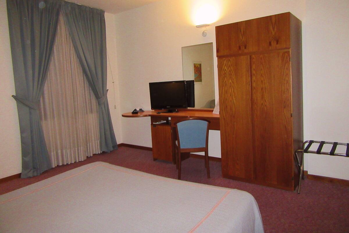 A hotel room with a bed, a desk with a computer, a chair, a wardrobe, and a television.